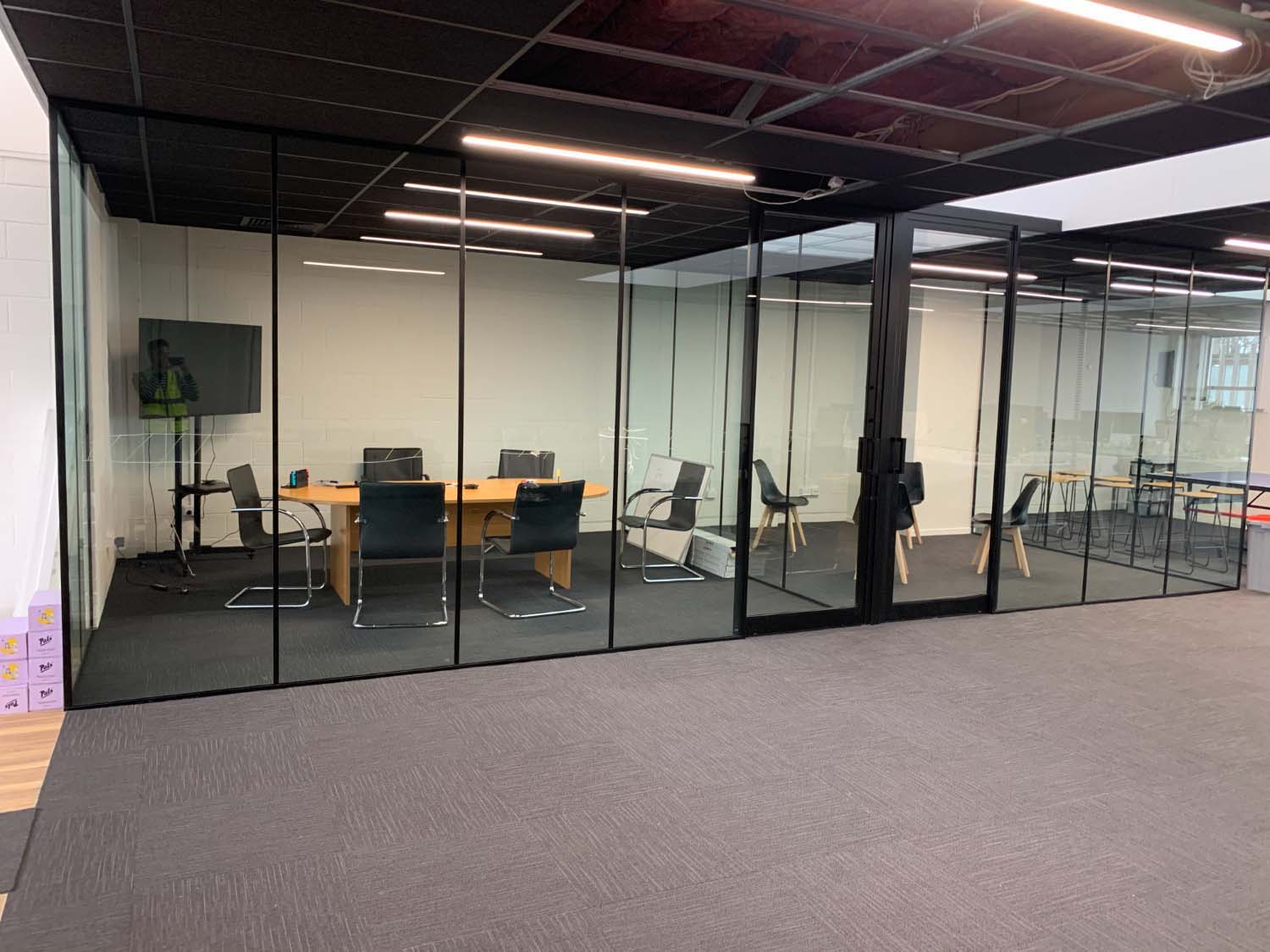 Black-framed glass partition meeting room with a wooden conference table, black cantilever chairs, and modern overhead strip lighting on dark carpet tiles.