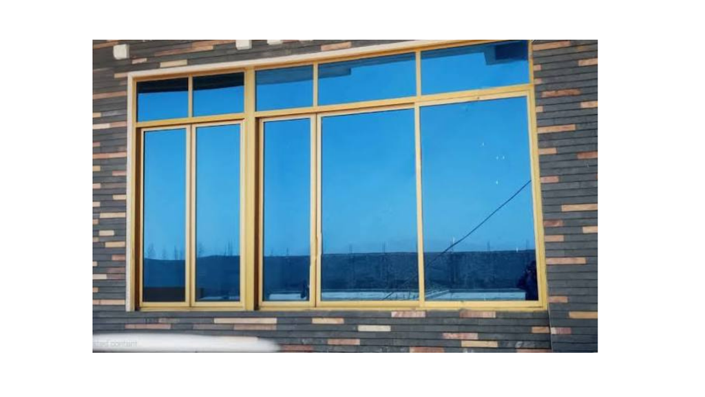 Sliding windows An architectural exterior window installation featuring heavy-duty aluminium profile frames in a metallic champagne/golden finish. The multi-section layout combines upper transom fixed glass panels with lower sliding and fixed units, paired with blue reflective heat-resistant glass installed against an exterior rustic brick tile facade.