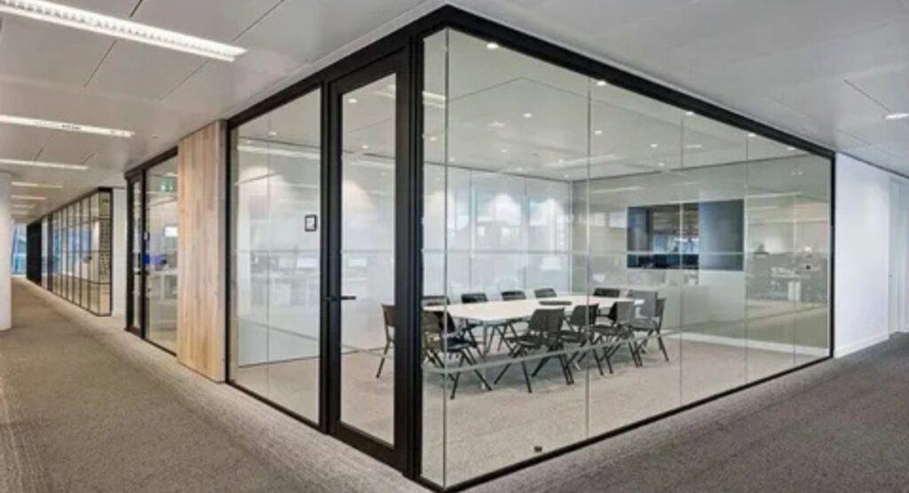 Corner view of a modern glass office partition enclosing a meeting room with a conference table, chairs, and a black aluminium door frame.