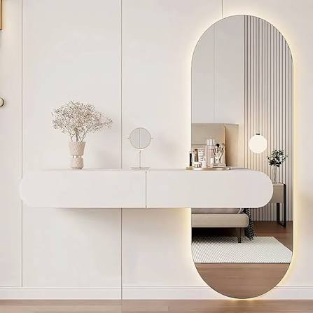 This high-resolution interior capture showcases a luxurious, classically inspired bathroom vanity setup highlighted by a tall, vertically mounted rectangular mirror. Encased in an intricately detailed, multi-tiered golden frame featuring fine beaded and fluted molding, the mirror serves as an elegant statement centerpiece against a soft, warm neutral wall.
Directly below, a rustic, textured natural limestone or travertine floating vanity slab supports a seamlessly carved, curved basin paired with minimalist, sleek brass Glass and furniture & mirror services in karachi plumbing fixtures. Flanking both sides of the mirror are delicate, modern ribbed glass wall sconces that cast balanced, glare-free ambient lighting across the wall. The mirror’s clean, distortion-free reflective surface captures a glimpse of an adjoining room filled with crisp white plantation shutters, neutral seating, and vibrant greenery, effectively bouncing daylight back into the room and creating an open, layered sense of space and tranquility.